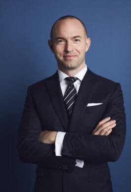 A person in a navy suit and striped tie stands against a solid blue background, smiling with crossed arms. 